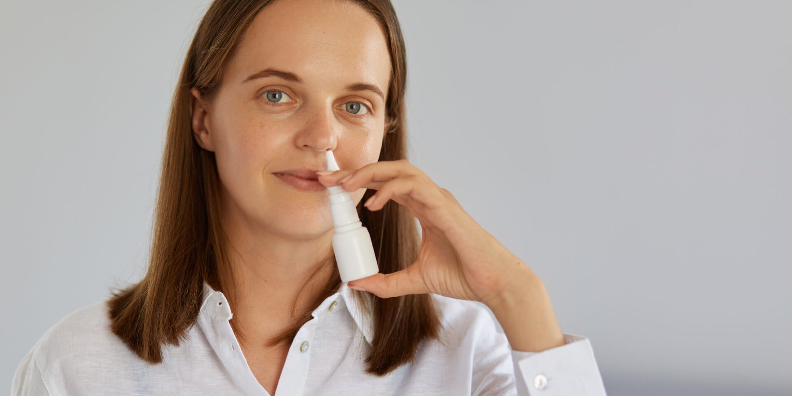 Closeup indoor shot of charming woman using nasal spray for runny nose, catches cold, looking at camera, wearing white shirt, posing against light wall.